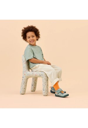 Lächendes Kind mit lockigem Haar sitzt seitlich auf einem gesprenkelten, klobigen Stuhl und trägt ein salbeigrünes Hemd, cremefarbene Hose, orangefarbene Socken und blaue Turnschuhe.