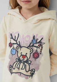 Kremowy hoodie z grafiką niedźwiedzia z czerwonym nosem, czarnym konturem oraz rogami ozdobionymi kolorowymi ornamentami i cukrową laską.