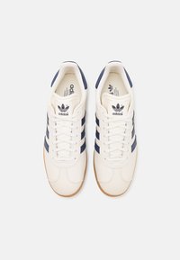 Adidas tenisky s krémovým koženým zvrškom, námorníckymi pruhmi a gumovou podrážkou. Obsahujú klasický dizajn na šnurovanie a logo na jazyku.