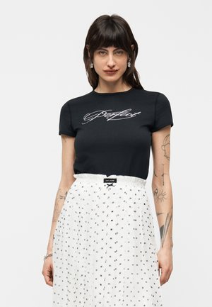 Kvinde med mørkt hår iført sort tætsiddende t-shirt med rhinstentekst og hvid højtaljet nederdel med lille sort firkantet mønster.