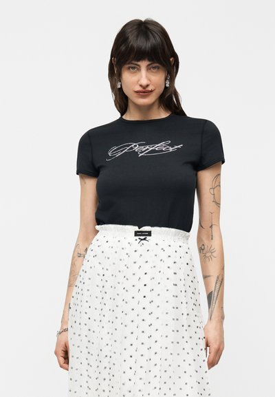 Femme aux cheveux foncés portant un t-shirt noir ajusté avec un texte en strass et une jupe blanche taille haute à motif de petits carrés noirs.