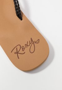 Ruskeat tasapohjaiset sandaalit, joissa on pehmeä teksturoitu pohja, musta punottu hihna ja tummanruskea kohokuvioitu "Roxy"-logo.