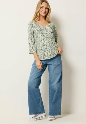 Lyshåret kvinde i en lys blomsterprintet bluse og brede blå jeans poserer med den ene hånd i lommen, iført hvide sneakers og smilende.