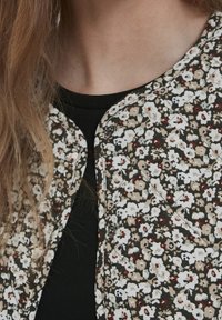 Zbliżenie na osobę w czarnej bluzce pod zapinaną kurtką w kwiatowy wzór z beżowymi, białymi i maroonowymi kwiatami.