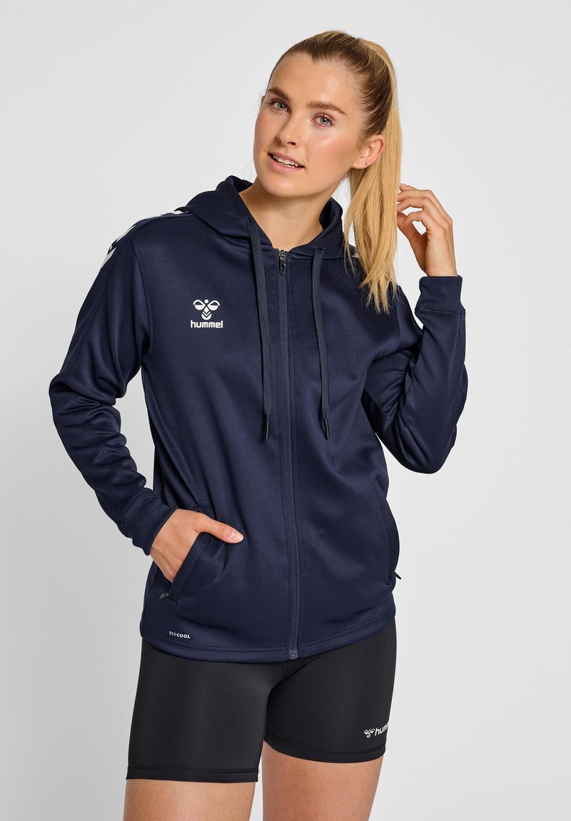 Junge Frau mit blondem Pferdeschwanz, die einen marineblauen Kapuzenpullover mit Reißverschluss und schwarze Shorts trägt, posiert mit einer Hand in der Tasche und der anderen nahe am Haar.