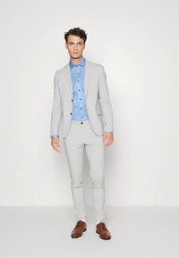 Abito grigio chiaro con vestibilità slim, abbinato a una camicia azzurra e scarpe in pelle marrone. Presenta un bavero a lancia e una texture sottile.