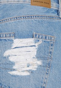 Jean en denim bleu clair avec une pièce usée sur la poche arrière. Les caractéristiques notables incluent une étiquette de marque en cuir et des bords effilochés.