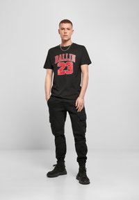 Juodas marškinėliai su raudona "BALLIN 23" grafika, derinami su juodomis krovinių kelnėmis. Modelis stovi su rankomis kišenėse, vilkėdamas juodus batus.