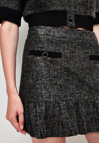 Falda de tweed negra y dorada con dobladillo acampanado, que presenta dos bolsillos de tela negra con acentos de anillo dorado y un acabado texturizado.