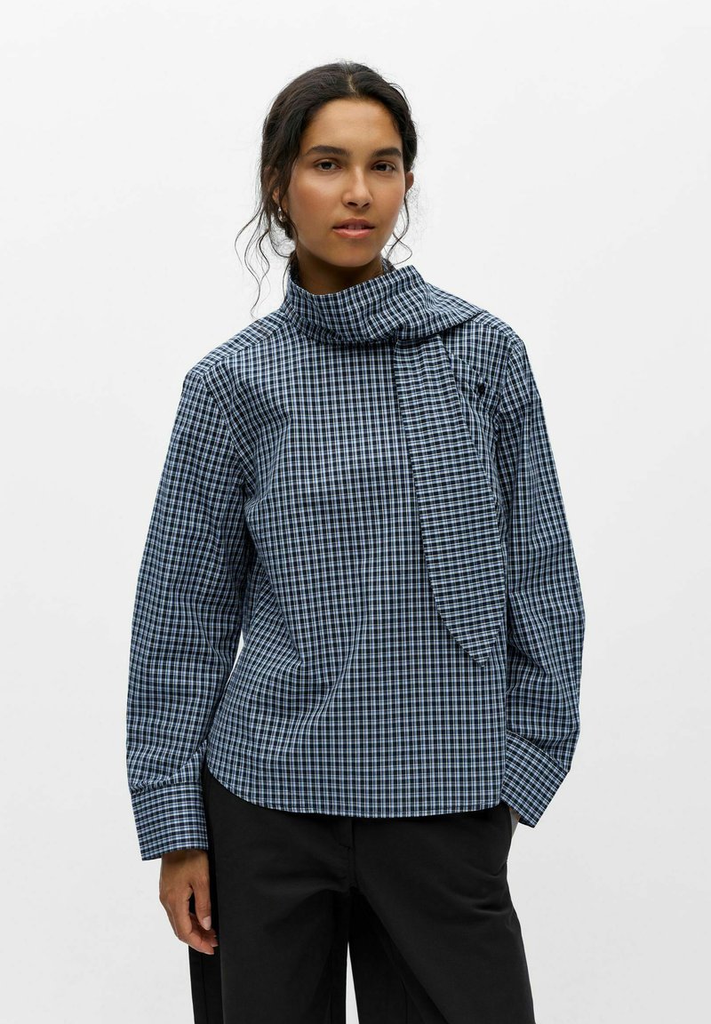 Femme aux cheveux foncés portant un chemisier à carreaux bleu et noir à manches longues avec un détail écharpe, associé à un pantalon noir, debout sur un fond blanc.