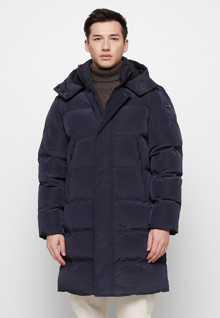 Donkerblauwe pufferjas met een ritssluiting aan de voorkant, twee zijzakken en een afneembare capuchon. Bevat een doorstikte design en een gladde, glanzende textuur.