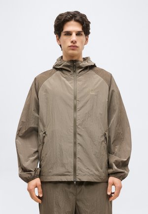 Junger Mann mit kurzen dunklen Haaren trägt eine hellbraune Kapuzen-Windjacke mit Frontreißverschluss und elastischen Bündchen, steht vor einfachem Hintergrund.