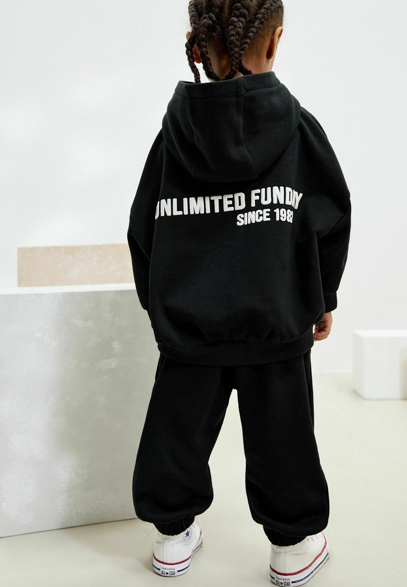 Kind mit geflochtenem Haar, das einen schwarzen Hoodie und schwarze Hosen trägt, weiße Converse-Sneaker, steht mit dem Rücken zur Kamera in einer minimalistischen Innenumgebung.