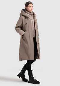 Dlouhý béžový péřový kabát s velkou kapucí, zipovými kapsami a elegantním designem. Nosený přes světlý top a v kombinaci s černými kotníkovými botami.