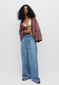 Model iført en løs, dyb maroon kimono med klokkeærmer over en lys gul bikini, parret med brede blå denim jeans.