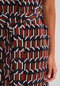 Robe à motifs multicolores présentant un design géométrique en rouge, marine, blanc et vert olive. Le tissu semble lisse avec un détail de taille nouée.