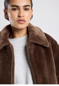 Ruskea tekoturkistakki, jossa on suuri kaulus, vetoketjusuljenta ja pehmeä pinta, päällä harmaa neulepaita. Kullanväriset renkaanmuotoiset korvakorut näkyvissä.