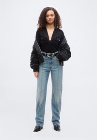 Γυναίκα με σγουρά μαλλιά που φοράει μαύρη μπλούζα με βαθιά V λαιμόκοψη, μαύρο μπουφάν bomber, τζιν ψηλόμεση μπλε με λεπτομέρειες στη ραφή και μαύρα μυτερά παπούτσια.