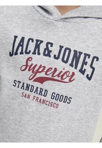 Felpa grigia realizzata in un tessuto dal tocco morbido. Presenta un audace testo stampato in blu navy e rosso: "JACK & JONES Superior Standard Goods SAN FRANCISCO."