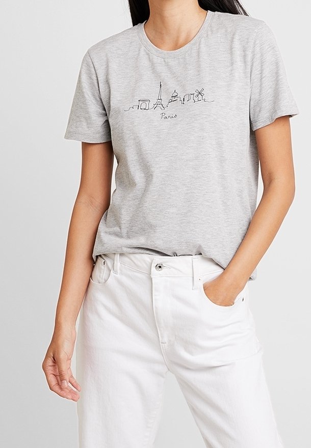 Femme portant un t-shirt gris clair avec un dessin au trait noir des monuments de Paris et un pantalon blanc taille haute, main dans la poche.