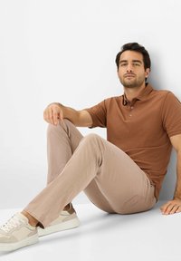 Bruine polo, lichtgekleurde, gestructureerde broek en lichtgekleurde sneakers met donkere accenten. De figuur zit tegen een effen achtergrond.