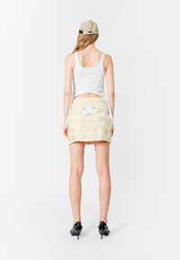 Top blanco sin mangas con espalda en forma de u, combinado con una falda corta beige que presenta tiras horizontales y un bolsillo de parche. Zapatos de tacón negros completan el look.