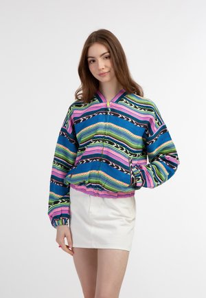 Kleurvolle ritsjack met een mix van blauwe, roze, groene en zwarte strepen en patronen; elastische manchetten en zoom; gecombineerd met een witte rok.
