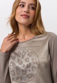 T-shirt beige à manches longues avec un tissu lisse, présentant un design en imprimé léopard circulaire en clous argentés et un col rond.