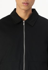Veste noire zippée avec un col, deux poches frontales et une fermeture éclair argentée ; texture lisse et design simple et minimaliste.
