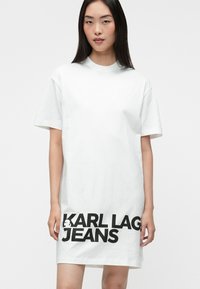 Balta kokvilnas T-kreklukleita ar īsām piedurknēm, apaļu kakla izgriezumu un lielu melnu tekstu "KARL LAGERFELD JEANS" apakšmalā.