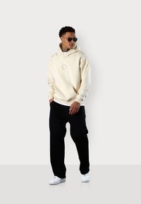 Cream hoodie with kangaroo pocket, featuring a small circular logo. Black loose-fitting pants and white sneakers complete the outfit.