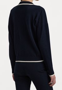 Henkilö, joka on pukeutunut tummansiniseen neuleeseen, jossa on valkoista reunusta kauluksessa, hihoissa ja helmassa, yhdistettynä samansävyisiin tummansinisiin housuihin, näkyy selkäpuolelta.