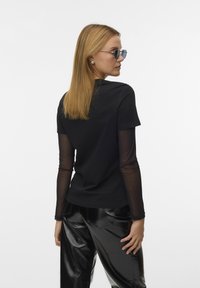 Top nero a maniche corte con maniche lunghe nere trasparenti; abbinato a pantaloni neri lucidi; la modella indossa occhiali da sole blu e ha capelli biondi lunghi e lisci.