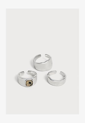 Trois bagues ouvertes en argent avec des designs lisses et modernes, dont une avec une petite pierre tachetée noire et beige.