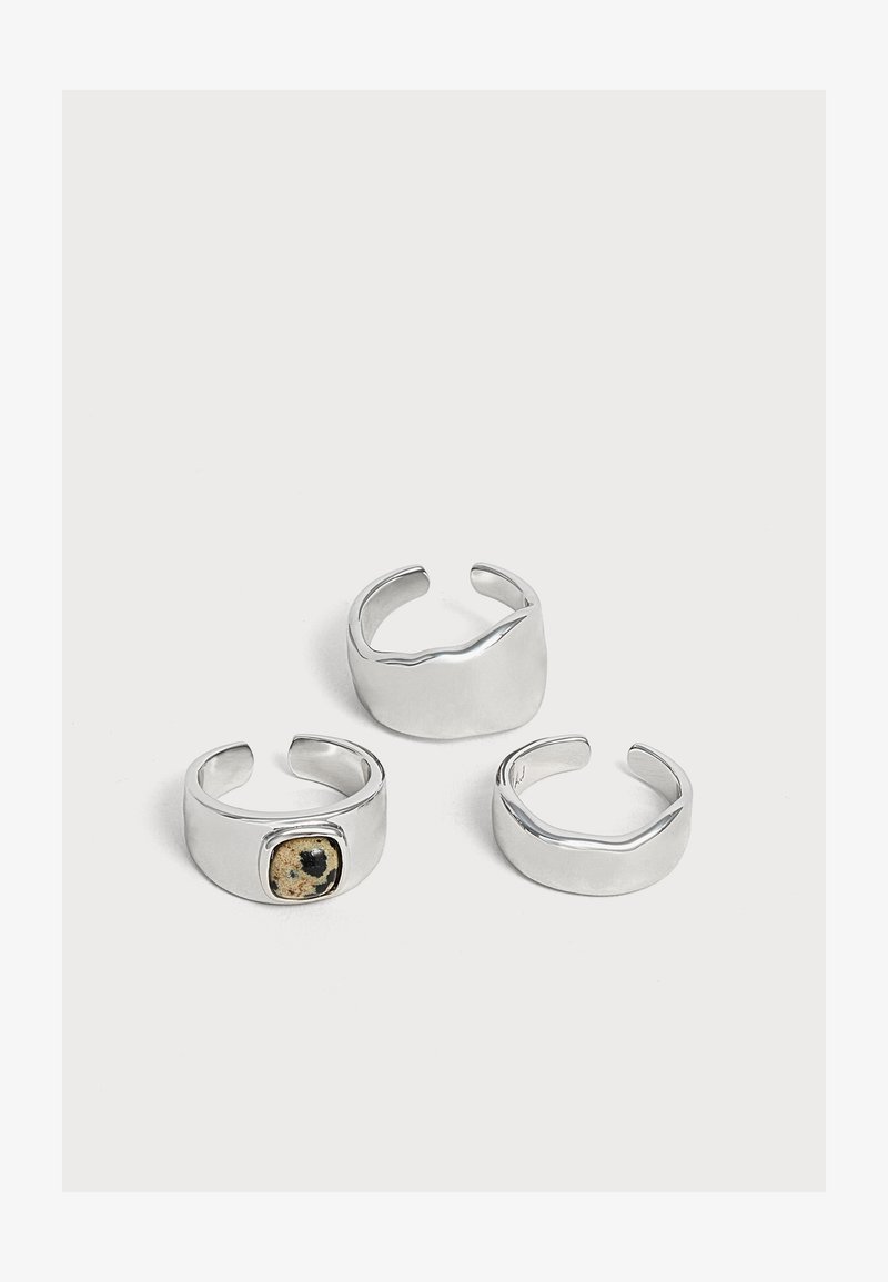 Trois bagues ouvertes en argent avec des designs lisses et modernes, dont une avec une petite pierre tachetée noire et beige.