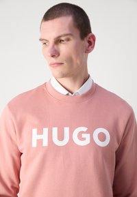Ružový sweatshirt s okrúhlym výstrihom, na prednej strane s veľkým bielym textom "HUGO"; vyrobený z mäkkej bavlny.