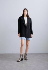 Woman standing against a plain wall wearing a black oversized blazer, white top, light denim shorts, and metallic heeled ankle boots.