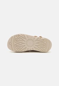 Sålen på en sko med et strukturert, lys beige gummimateriale med et sollys-mønster og merkevarelogo, designet for grep og holdbarhet.