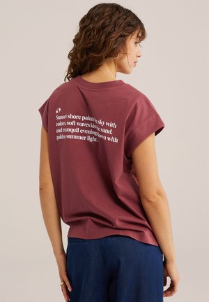 Žena s kučeravými vlasmi, ktorá nosí bordové tričko s bielym textom na chrbte, v kombinácii s modrými džínami, stojaca na jednofarebnom pozadí.