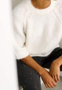 Personne assise portant un pull en maille blanche texturée à manches trois-quarts et un pantalon noir orné de petits points blancs.