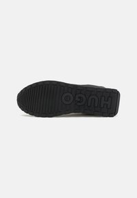Semelle de sneaker en caoutchouc noir présentant un motif texturé avec des rainures horizontales et verticales, et le logo "HUGO" embossé sur le côté.