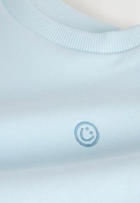 Tissu bleu clair avec un petit visage souriant bleu brodé près du col côtelé d'un vêtement.