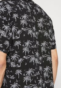 Lyhythihainen musta paita, jossa valkoiset palmu- ja purjelaivakuviot, rento istuvuus ja napillinen kaulussuunnittelu.