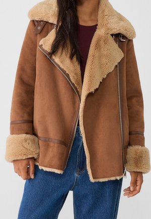 Person wearing a brown shearling jacket with large lapels, zipper closure, and fur cuffs over a burgundy top and blue jeans.