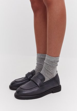 Black leather loafers with a round toe and a front strap, paired with folded gray socks, on bare legs against a light background.