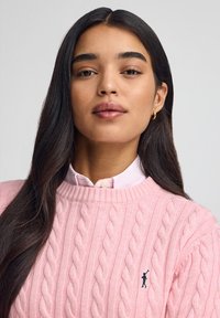 Ung kvinna med långt mörkt hår, klädd i en rosa kabelstickad tröja och guldfärgade ringörhängen, poserande framför en enfärgad bakgrund.