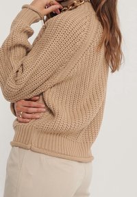 Beige pletený svetr s vysokým límcem, hřebínkovými manžetami a spodním lemem; má strukturovaný vzor a volný střih. Doplněno s světlými kalhotami.