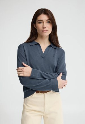 Donna con capelli castani lunghi che indossa un maglione polo blu a maniche lunghe e pantaloni crema con una cintura marrone, in posa con le braccia incrociate.