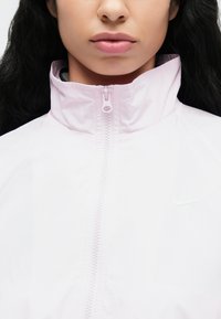 Το ανοιχτό ροζ μπουφάν με φερμουάρ διαθέτει ψηλό γιακά, λείο ύφασμα και ένα μικρό λογότυπο της Nike στο πλάι. Το φερμουάρ κλεισίματος βρίσκεται στο κέντρο.