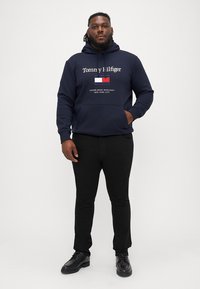 Sötétkék kapucnis pulóver "Tommy Hilfiger" felirattal és zászló mintával az elején. Fekete nadrággal és fekete cipővel párosítva, ami egy laza öltözetet alkot.